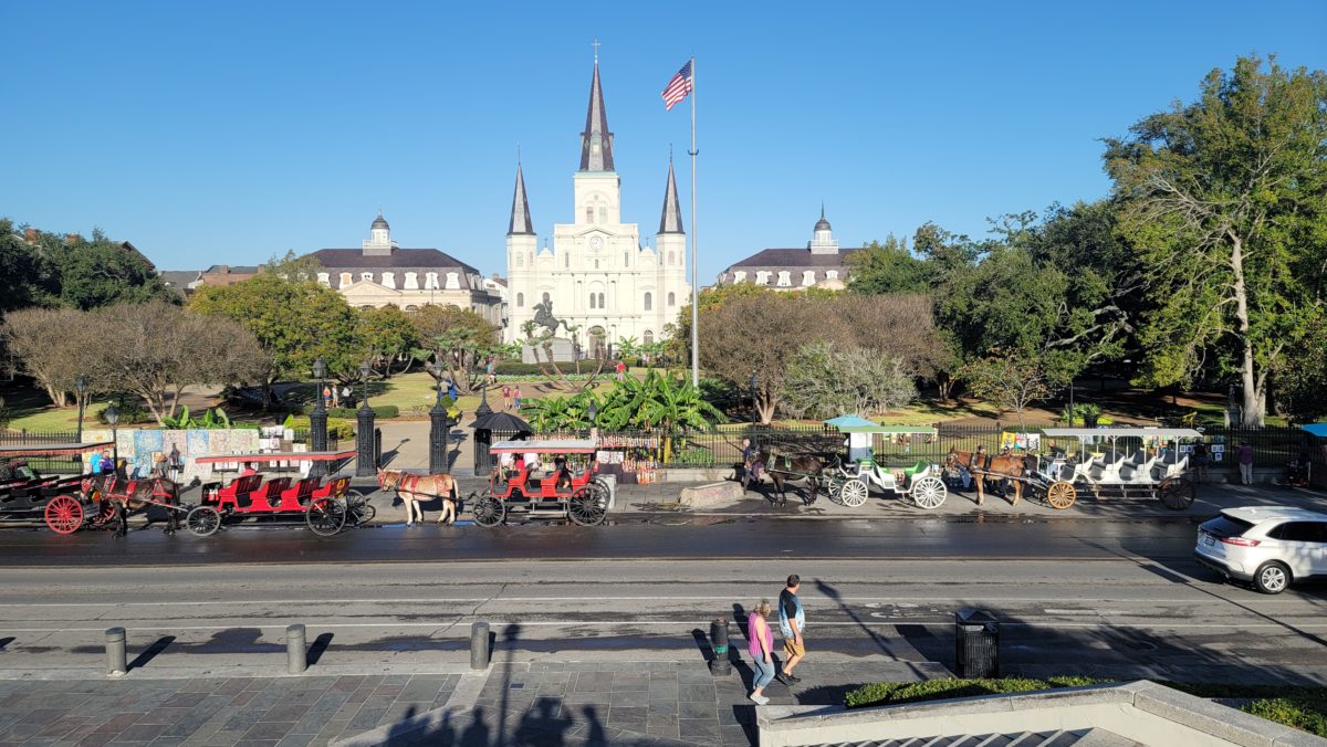 In the background is a c=white cathedral with a black roof. The mid ground is Jackson Park. In the foreground is line of horse drawn buggys waiting for customers