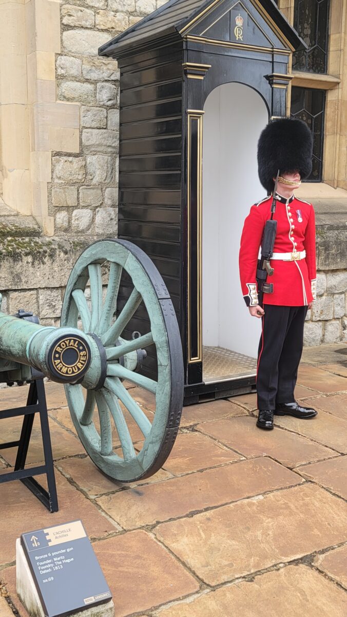 A British guard in the traditional red uniform with a tall bear skin hat in front of a guard house, next to an old cannon. In front of the Tower of London