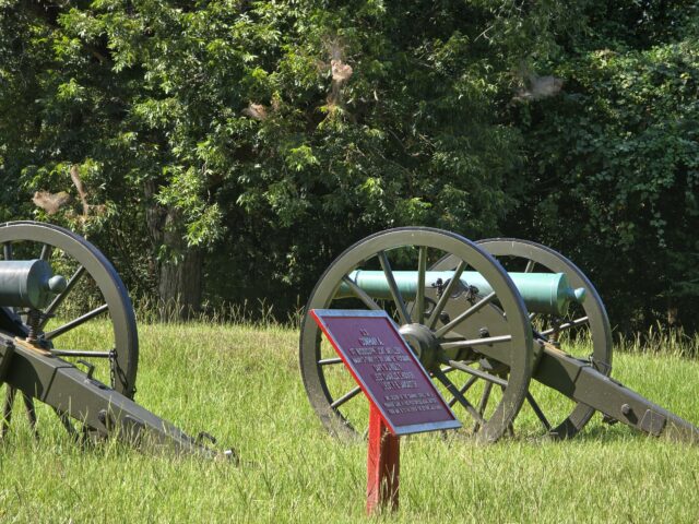 two bronze cannon on a grass field near a dense forest. There is a sign indicating the cannons were a gun emplacement for the Confederate Army